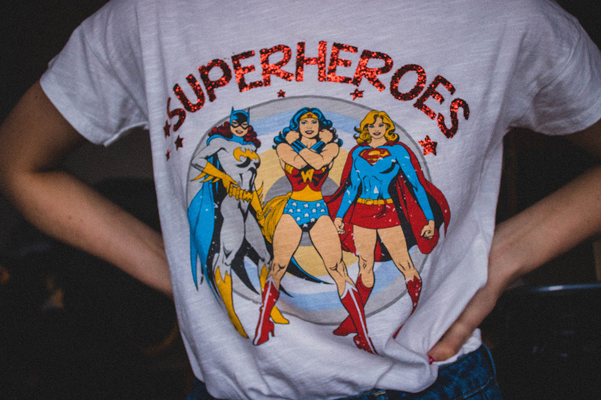 A woman with showcasing a t-shirt that says Superheroes in sparkly font, with DC female heroes pictured beneath the words.